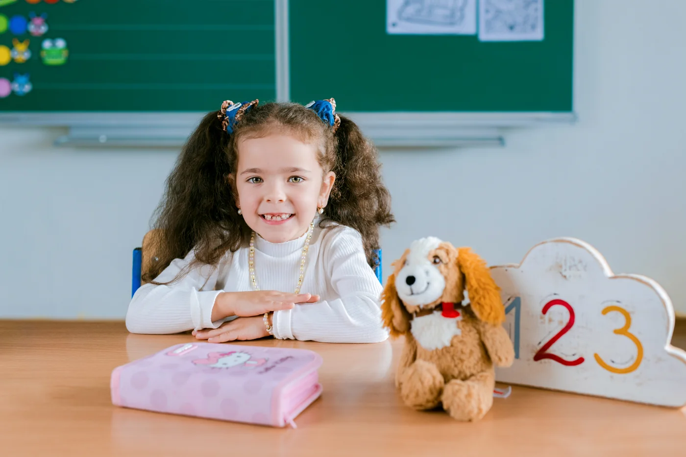Usměvavá prvňačka sedí ve třídě u lavice s penálem a hračkou